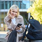 lifestyle-portrait-smiling-female-model-sitting-street-bench-with-backpack-mobile-phone